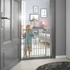 A toddler stands behind a silver baby gate in a doorway, looking into a room with furniture and artwork. Voir les images du produit