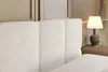 A close-up view of a beige upholstered headboard of a bed, with a bedside table and lamp visible on the right. Produktbild-Vorschau