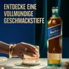 A hand holding a glass of Johnnie Walker Blue Label whisky next to the bottle, with promotional text in German. Produktbild-Vorschau
