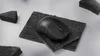 A black wireless mouse is placed on two dark textured square platforms, surrounded by dark textured triangular blocks. Produktbild-Vorschau