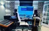 A recording studio setup with acoustic panels, a computer monitor displaying audio software, a mixing board, a keyboard, a chair, and two electric guitars. Podgląd zdjęcia produktu