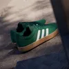 A pair of green Adidas sneakers with white stripes and a gum sole are placed on a concrete surface, with shadows cast by sunlight. Produktbild-Vorschau