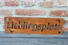 A rusty metal sign with the word "Lieblingsplatz" cut out, placed against a brick wall. Produktbild-Vorschau