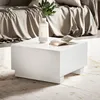 A white high-gloss coffee table is shown in a living room setting, with a draped sofa in the background and a rug on the floor. A vase with a plant and an open book are on the table. Produktbild-Vorschau