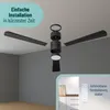 An exploded view of a ceiling fan with its components disassembled, mounted on a white ceiling, with a view of a kitchen area in the bottom right corner showing a remote control mounted on the wall. Produktbild-Vorschau