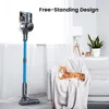 A cordless vacuum cleaner is shown standing upright next to a sofa where a cat is resting. The scene is set in a modern living room with a window and a lamp in the background. Produktbild-Vorschau