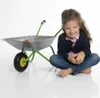 A young girl sits cross-legged next to a child-sized wheelbarrow on a white background. Produktbild-Vorschau