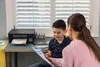 A mother and son are sitting at a desk with a printer, looking at a document. Podgląd zdjęcia produktu