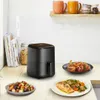 A black air fryer is shown on a kitchen counter with plates of food, books, and mugs in the background. Produktbild-Vorschau