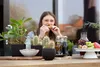 A woman is eating at a table with plants and fruit. Produktbild-Vorschau