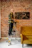 A man stands on a ladder in front of a brick wall, next to a yellow sofa. Anteprima dell’immagine prodotto