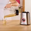 A person is pouring a smoothie from a blender pitcher into a glass, with the blender base visible on a wooden table. Produktbild-Vorschau