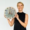 A woman is holding a round wall clock with a sailboat painting on it. Produktbild-Vorschau
