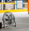 A floor fan is positioned on a textured floor in what appears to be a workshop or industrial setting, with yellow and blue shelving visible in the background. Náhľad obrázku produktu