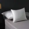 Two white silk pillowcases are placed on a gray bed. A bedside table with a lamp is visible on the left. Podgląd zdjęcia produktu