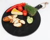 A round black serving board with various food items arranged on it, including sliced pretzel bread, cheese, tomatoes, red pepper, cucumber slices, and microgreens. Anteprima dell’immagine prodotto