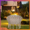A hanging chair is suspended in a garden at dusk, with a house and illuminated trees in the background. Voir les images du produit
