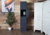 A tall, dark gray metal locker with three compartments stands in a room with a wooden wall and a white folding screen. A potted plant is to the left, and a painting of a woman's face is on the wall. Produktbild-Vorschau