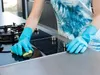 A person wearing blue rubber gloves is cleaning a modern kitchen stovetop with a yellow sponge. Náhľad obrázku produktu