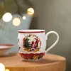 A Christmas-themed mug sits on a wooden table, with blurred festive lights in the background. Voir les images du produit