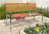 A wooden and metal bench with a blanket and book on it, placed on a patio with flowers. Produktbild-Vorschau