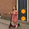 A young girl is smiling while standing on a pink electric scooter on a sidewalk in front of a brick wall and a dark blue door. Anteprima dell’immagine prodotto