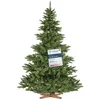 A green artificial Christmas tree with a wooden stand, featuring a tag with product information. Produktbild-Vorschau