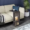 A dark gray side table with vertical wooden slats is placed next to a beige sofa in a living room setting. A tablet and a cup of coffee are on the table, and a book is in the lower shelf. A blanket is draped over the sofa, and a potted plant is visible in  Produktbild-Vorschau