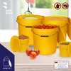 Several yellow buckets filled with different items, including liquid, grain, tomatoes, and kibble, are arranged on a table. Three red apples are in the foreground. The background is a bright, indoor setting with a window. Voir les images du produit