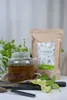 A glass teapot filled with herbal tea, a bag of dried herbs, and fresh mint and lime slices are arranged on a dark surface, with a blurred background of greenery. Produktbild-Vorschau