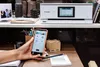 A Canon PIXMA TS8751 printer is shown on a shelf above a desk, with a smartphone in the foreground displaying the Canon app. Podgląd zdjęcia produktu