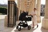 A woman pushing a black stroller along a colonnade. Produktbild-Vorschau
