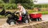 A young boy is riding a toy tractor through a muddy field, pulling a red trailer filled with hay. Produktbild-Vorschau