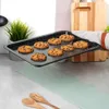 A baking tray with chocolate chip cookies is being held by a hand wearing an oven mitt, with kitchen utensils and a plant in the background. Voir les images du produit