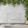 A white metal garden bench is placed on a stone patio in front of a white wall, with green vines and white flowers arching overhead. Podgląd zdjęcia produktu