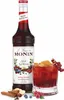 A bottle of Monin Spiced Red Berries syrup next to a glass of red beverage, with some ingredients like cinnamon sticks and ginger in the foreground. Voir les images du produit
