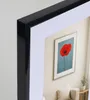 A framed picture of a red poppy on a blue background, displayed in a living room setting with a sofa and a small plant. Produktbild-Vorschau