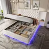 A white upholstered bed with a tufted headboard is shown with its storage compartment open, revealing various neatly folded items. Blue LED lights are visible underneath the bed frame. Náhled obrázku produktu