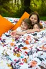 Three children are lying on a bed with a floral and bird-patterned duvet and orange pillows, outdoors in a garden setting. Produktbild-Vorschau