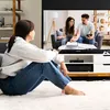 A young woman sits on a fluffy white rug, holding a remote control and watching a TV screen. The TV displays a couple arguing over documents. Voir les images du produit