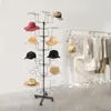 A black metal hat stand with multiple tiers displaying various hats, set against a white and light gray background. In the background, a clothing rack with garments is visible. Produktbild-Vorschau
