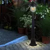 A black outdoor lamp post with a glowing bulb stands on a cobblestone path, surrounded by potted plants and a rustic wooden cart. Produktbild-Vorschau