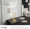 A modern table lamp with a gold base and white rectangular shade is displayed on a dark dresser, next to a piece of abstract art and decorative objects. Text overlays describe its features. Produktbild-Vorschau