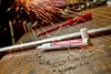 A paint marker and a screwdriver are on a wooden surface with sparks flying in the background. Produktbild-Vorschau