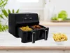 A dual-basket air fryer filled with fries and chicken wings, placed on a wooden table with a bowl of fries in the foreground. The background shows a blurred kitchen setting with plants and a head of lettuce. Voir les images du produit