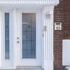 A white front door with a glass pane and a side window, next to a brick wall with a house number sign. Podgląd zdjęcia produktu