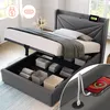 A grey upholstered bed frame with the mattress lifted to reveal storage compartments underneath. A phone is shown charging on the headboard. Produktbild-Vorschau
