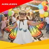 A young girl dressed as a butterfly is smiling and posing for a photo at an outdoor event with balloons and other people in the background. Produktbild-Vorschau
