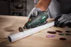 A person wearing gloves is using a cordless rotary tool to cut a white pipe on a wooden table. Several cutting discs are scattered around the pipe. Produktbild-Vorschau
