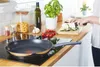 A person is preparing food in a kitchen, with a frying pan on the stove and vegetables being chopped. Voir les images du produit